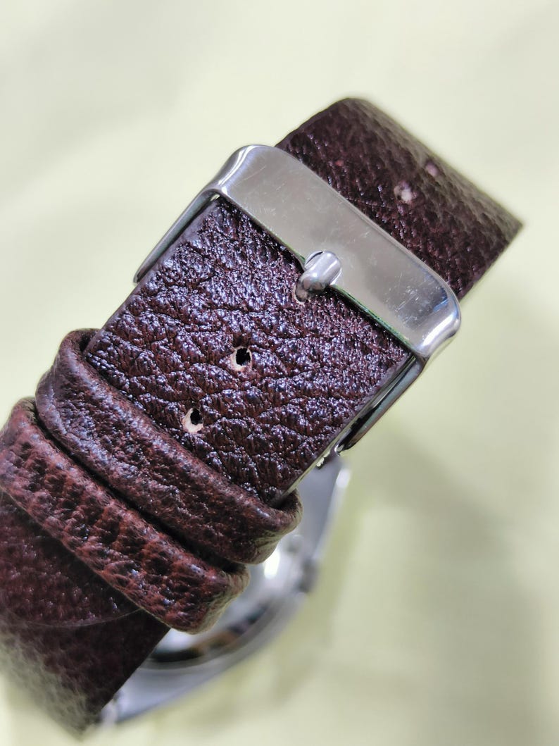 May include: Close-up of a brown leather watch strap with a silver buckle. The strap has a textured surface and is secured with a metal clasp. The watch face is partially visible.