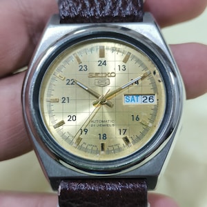 May include: A vintage Seiko wristwatch with a gold-toned face and a stainless steel case. The watch features a brown leather strap with white stitching. The face displays the day and date, with the day reading "SAT 26". The watch is marked "AUTOMATIC 21 JEWELS".