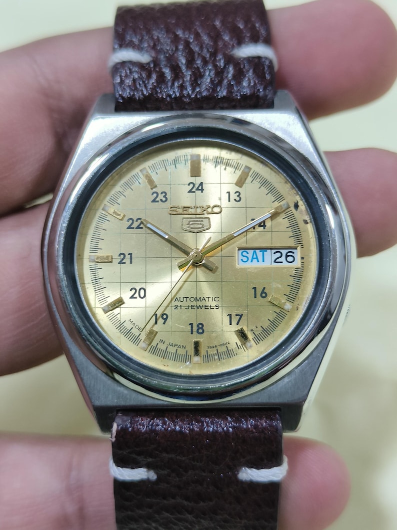 May include: A vintage Seiko wristwatch with a gold-toned face and a stainless steel case. The watch features a brown leather strap with white stitching. The face displays the day and date, with the day reading "SAT 26". The watch is marked "AUTOMATIC 21 JEWELS".