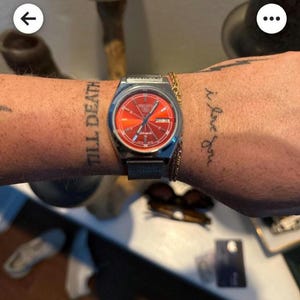 Könnte beinhalten: Eine silberne Armbanduhr mit rotem Zifferblatt und Mesh-Armband wird gezeigt. Die Uhr befindet sich an einem Handgelenk mit dem Tattoo "TILL DEATH". Ein goldenes Kettenarmband ist ebenfalls vorhanden.