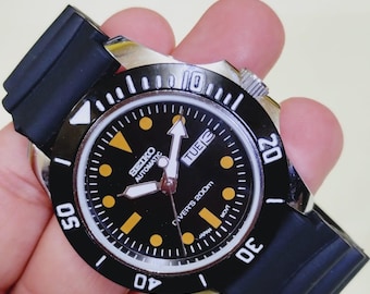 May include: Black and silver Seiko automatic diver's watch with a black dial, yellow hour markers, and a black rubber strap. The watch face displays the day of the week and is water resistant to 200m.