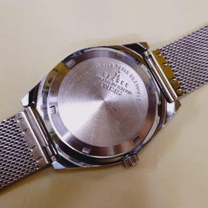 K&ouml;nnte beinhalten: Eine Seiko-Uhr aus Edelstahl mit einem Mesh-Armband. Der Uhrenr&uuml;cken ist sichtbar und zeigt das Seiko-Logo, die Modellnummer 335718 sowie die Worte "Water Resistant" und "Stainless Steel".