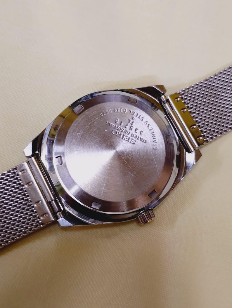 K&ouml;nnte beinhalten: Eine Seiko-Uhr aus Edelstahl mit einem Mesh-Armband. Der Uhrenr&uuml;cken ist sichtbar und zeigt das Seiko-Logo, die Modellnummer 335718 sowie die Worte "Water Resistant" und "Stainless Steel".