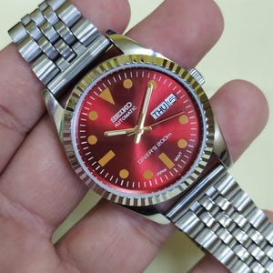 Puede incluir: Un reloj de pulsera automático Seiko de acero inoxidable con esfera roja. El reloj presenta marcadores de hora y agujas doradas, una pantalla de día y fecha y un bisel texturizado. La correa del reloj es de acero inoxidable.