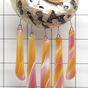 Op de afbeelding: Een decoratieve windgong met een grote, iriserende zeeschelp met een spiraalvormig ontwerp. Vijf langwerpige glazen hangers in tinten geel, oranje en roze hangen eronder, waardoor een kleurrijk en opvallend display ontstaat. De windgong is opgehangen aan dunne rode koorden.