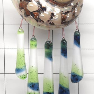 May include: A decorative wind chime featuring a large, iridescent brown and cream-colored seashell at the top. Five elongated, teardrop-shaped glass pieces in shades of white, lime green, and deep blue hang from the shell by thin copper wires.