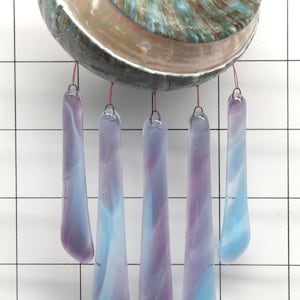 Peut inclure: Un carillon décoratif avec un grand coquillage irisé aux tons turquoise, marron et crème. Cinq pièces de verre allongées dans les tons bleus et violets pendent en dessous, créant une esthétique apaisante.