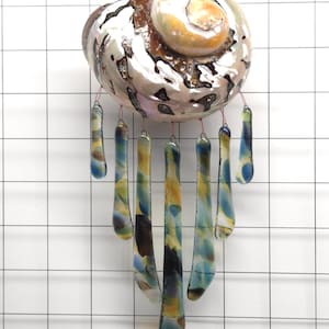 Op de afbeelding: Een decoratieve windgong met een grote, iriserende zeeschelp met een spiraalvormig ontwerp. Zeven glazen hangers in blauwe, gele en bruine tinten hangen aan de schelp en creëren een uniek en artistiek woondecoratie-item.