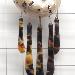 Op de afbeelding: Een decoratieve windgong met een grote, iriserende zeeschelp met een spiraalvormig ontwerp. Vijf langwerpige glazen hangers in tinten amber, bruin en blauw hangen eronder, opgehangen aan rode koorden. De achtergrond is een wit raster.