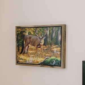May include: A framed artwork depicting a deer in a forest setting hangs on a white wall. A round mirror with a wooden frame is visible. A small wooden side table holds a book and a potted plant. Decorative pillows rest on a bed.