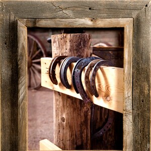 May include: A series of five dark metal horseshoes hanging over a horizontal light-colored wooden beam, which is attached to a vertical wooden post. The entire scene is enclosed within a weathered, dark brown wooden picture frame.
