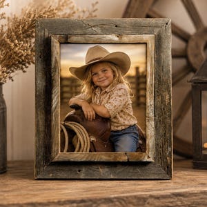 May include: A rectangular picture frame crafted from weathered, dark brown reclaimed wood with a visible grain. The frame holds a photograph of a child wearing a tan cowboy hat, a light-colored patterned shirt, and blue jeans, leaning on a leather saddle.