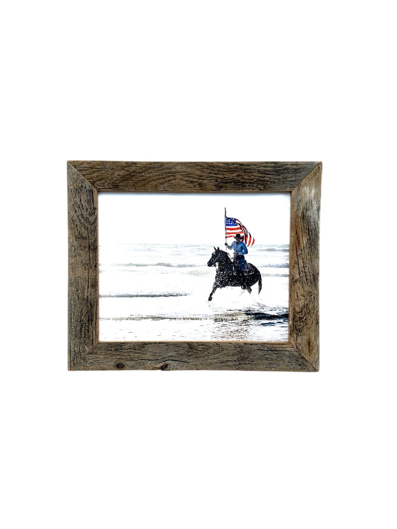 May include: A framed print featuring a person riding a black horse through water, holding a large American flag. The frame is made of rustic, weathered wood. The image is set against a white background.