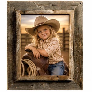 May include: A rectangular picture frame crafted from weathered, dark brown reclaimed wood with a visible grain. The frame encloses a photograph of a child wearing a tan cowboy hat and a patterned shirt, leaning on a brown leather saddle.
