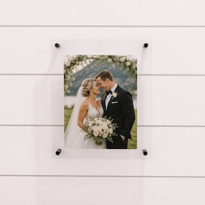 May include: A framed photograph of a wedding couple, mounted on a white wall. The photo shows a bride and groom embracing under a floral arch. The frame is clear acrylic, held in place by four black fasteners.
