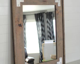 May include: A rectangular mirror with a rustic wooden frame and silver metal corner accents. The mirror is hanging on a white wall.