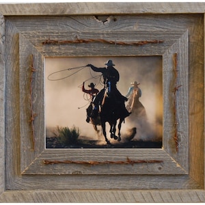 May include: A rustic wooden picture frame with barbed wire accents, featuring a photograph of three cowboys on horseback riding through a dusty field.