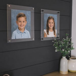 May include: Two clear acrylic photo frames mounted on a dark gray wall. Each frame holds a portrait of a smiling child. A vase with white flowers and a cream-colored vase with a yellow base sit on a wooden surface.