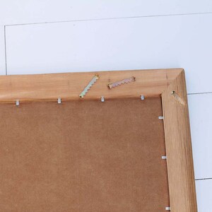 May include: A close-up of the back of a wooden picture frame with metal hanging hardware attached. The frame is made of natural wood with a brown stain. The back of the frame is made of brown cardboard.