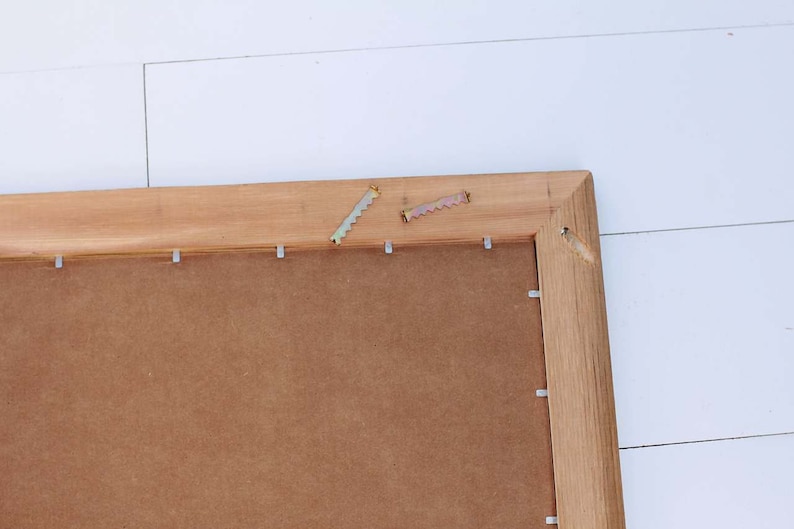 May include: A close-up of the back of a wooden picture frame with metal hanging hardware attached. The frame is made of natural wood with a brown stain. The back of the frame is made of brown cardboard.