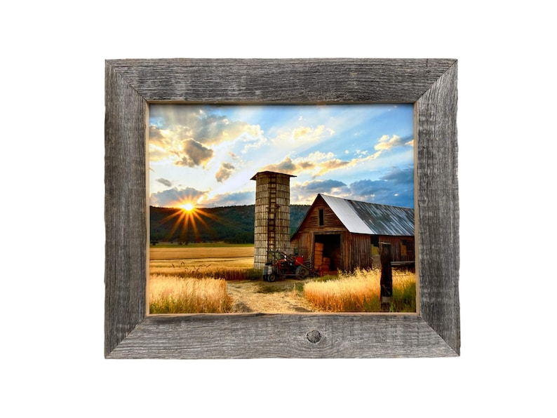 May include: A framed print of a rural landscape featuring a barn, silo, and tractor at sunset. The wooden frame is gray. The sky is filled with clouds and the sun is setting over a field of wheat.