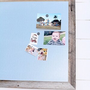 May include: A light blue magnetic board with a wood frame, holding several photos of people. The photos are held in place with small black magnets.