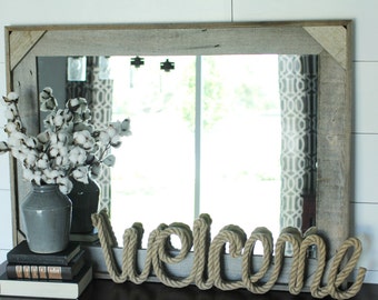 May include: A rustic wooden framed mirror with a rope welcome sign in front of it. A vase with white cotton flowers sits on a stack of books to the left of the sign.