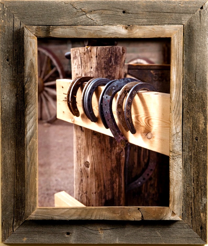May include: A series of five dark metal horseshoes hanging over a horizontal light-colored wooden beam, which is attached to a vertical wooden post. The entire scene is enclosed within a weathered, dark brown wooden picture frame.