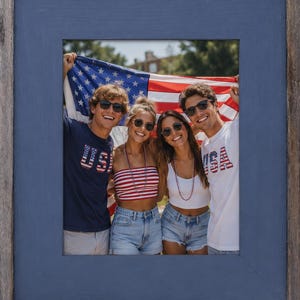 May include: A framed photograph featuring four people holding an American flag. The frame is a weathered blue color. The people are wearing sunglasses and patriotic clothing, including shirts with "USA" designs and red, white, and blue striped tops.