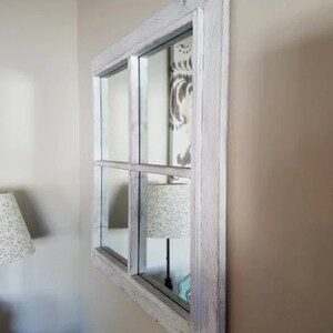 Puede incluir: Un espejo con marco de madera blanqueado con un dise&ntilde;o de ventana de cuatro paneles. El espejo est&aacute; colgado en una pared de color beige claro.
