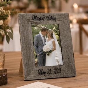 May include: A rectangular picture frame made of weathered gray wood with a rough texture. The top of the frame is engraved with the text "Mitch & Ashley" and the bottom is engraved with "May 21, 2011". It holds a photograph of a couple.