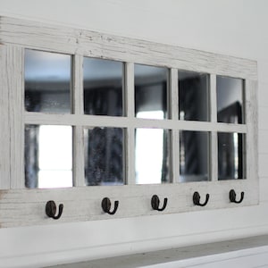 May include: Whitewashed wood framed mirror with nine panes and five hooks underneath. The mirror is hanging on a white wall.