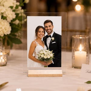 May include: A clear acrylic photo frame with a wooden base displays a wedding photo of a smiling couple. The table setting includes a lit candle in a glass holder, white flowers, and a table number card.