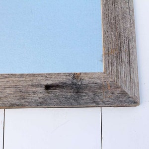 May include: Close-up of the corner of a rustic wooden frame with a light blue background. The frame is made of weathered wood with knots and grain.