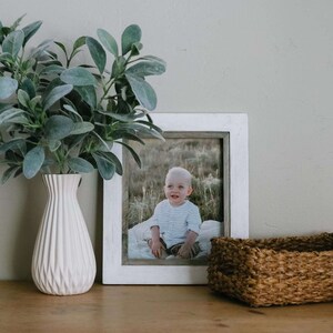 May include: A white picture frame with a photo of a young child sitting on a blanket in a field. The frame is on a wooden table next to a white vase with green plants and a brown wicker basket.