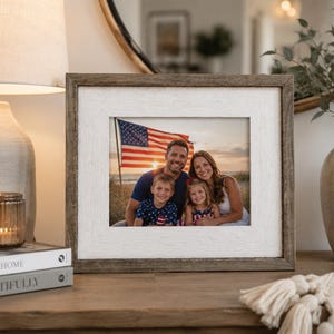 May include: A rectangular picture frame with a rustic brown wood outer border and a textured white inner mat. The frame contains a photograph of a family of four posing outdoors in front of an American flag during a sunset at the beach.