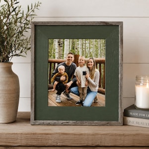 May include: A square picture frame with a weathered gray wood outer border and a dark forest green inner mat. The frame contains a photograph of two adults and two children posing together on a wooden deck in front of a forest of birch trees.