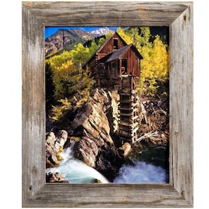 May include: A framed print of a rustic wooden mill perched on a rocky cliff, with a waterfall cascading below. The surrounding landscape features vibrant yellow and green trees, and a mountain range in the background. The frame is made of weathered wood.