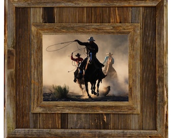 May include: A framed photograph of cowboys on horseback roping cattle. The image is set in a rustic wooden frame. The cowboys are wearing hats and riding horses in a dusty environment.