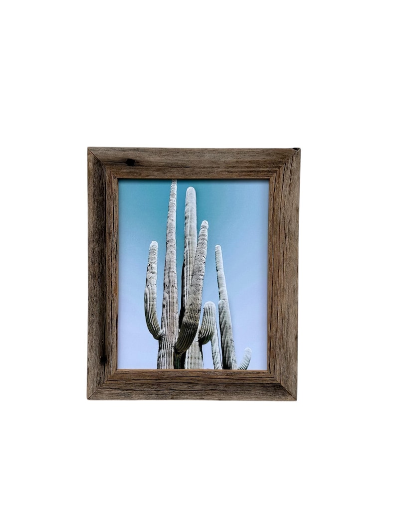 May include: Square framed print featuring a desert landscape with tall saguaro cacti against a light blue sky. The frame is made of weathered, light brown wood, adding a rustic touch to the artwork. The image evokes a sense of the American Southwest.