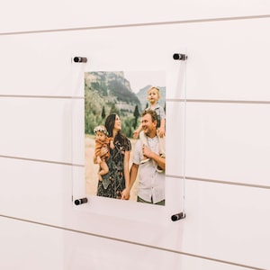 May include: A framed photo of a family of four in a mountain setting. The parents are holding their two children, one on each shoulder. The photo is mounted on a white wall with black clips.