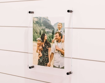 May include: A framed photo of a family of four in a mountain setting. The parents are holding their two children, one on each shoulder. The photo is mounted on a white wall with black clips.