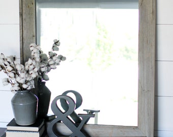 May include: A rustic wooden framed mirror with a gray wash finish sits on a dark wood table with a stack of books and a gray ceramic vase with cotton stems. A large gray wooden ampersand sits in front of the vase.