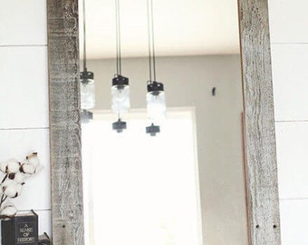 May include: A large rectangular mirror with a rustic, weathered wood frame. The frame is a light gray color with dark brown wood grain showing through. The mirror is reflecting a room with a white ceiling and a few hanging light fixtures.