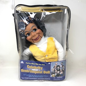 May include: A Celebrity Ventriloquist Doll in a clear plastic case. The doll has a smiling face, round glasses, and a yellow vest over a white turtleneck. The case has a black zipper and a blue label with text.