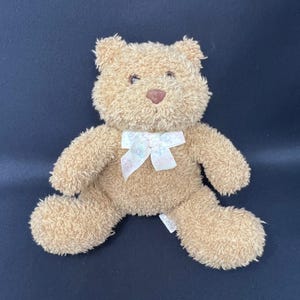 Puede incluir: Un oso de peluche con pelaje marrón claro, una nariz marrón y un lazo blanco con un estampado floral. El oso está sentado sobre una superficie azul oscuro.