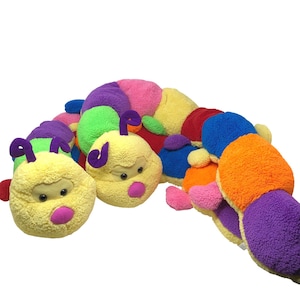 Pode incluir: Um brinquedo de pelúcia de lagarta multicolorida com um corpo longo e segmentado feito de tecido macio e felpudo. O brinquedo exibe uma gama vibrante de cores, incluindo amarelo, roxo, rosa, verde, azul e laranja, com duas cabeças.