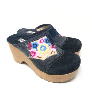 May include: A pair of platform clogs with a wooden sole and wedge heel. The upper is a combination of navy blue suede and denim, with a floral embroidered patch in pink, yellow, and blue. The clogs have a slip-on design.