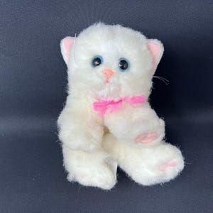 Puede incluir: Un juguete de peluche blanco con forma de gatito con ojos azules, nariz rosa y lazo rosa. El gatito tiene detalles rosas en las orejas y las patas. El juguete está hecho de material suave y esponjoso.