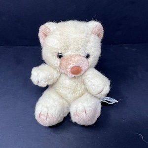 May include: A small, white plush teddy bear with pink accents on the ears, nose, and paws. The bear is sitting with its legs crossed and has a sweet expression.
